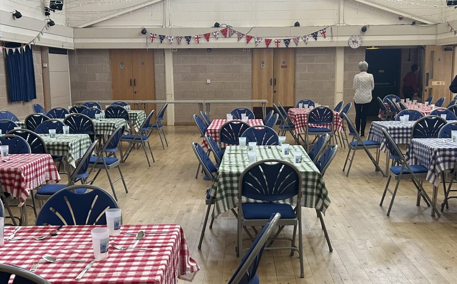 Tables with gingham table cloths and blue chairs set up for dinner in a dining hall with a wooden floor